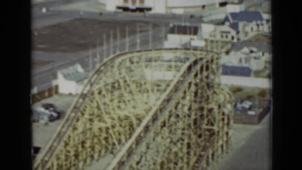 Elevated 1937 San Francisco view of the wooden roller coaster beside small amusement-park buildings, open pavement, and parked cars.