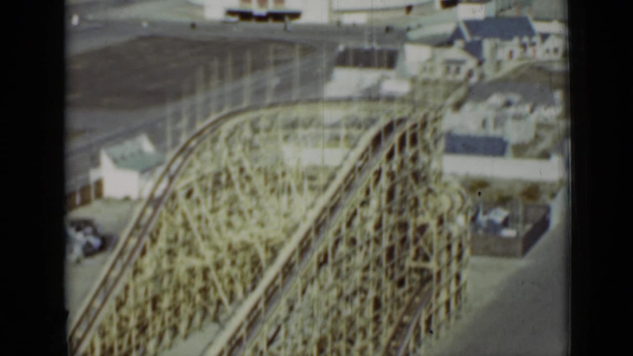 Wooden roller coaster structure seen from above near waterfront buildings.