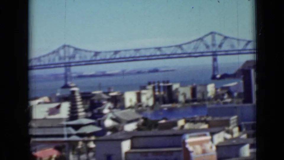 1937 San Francisco view of a steel truss bridge crossing the bay beyond low waterfront rooftops.