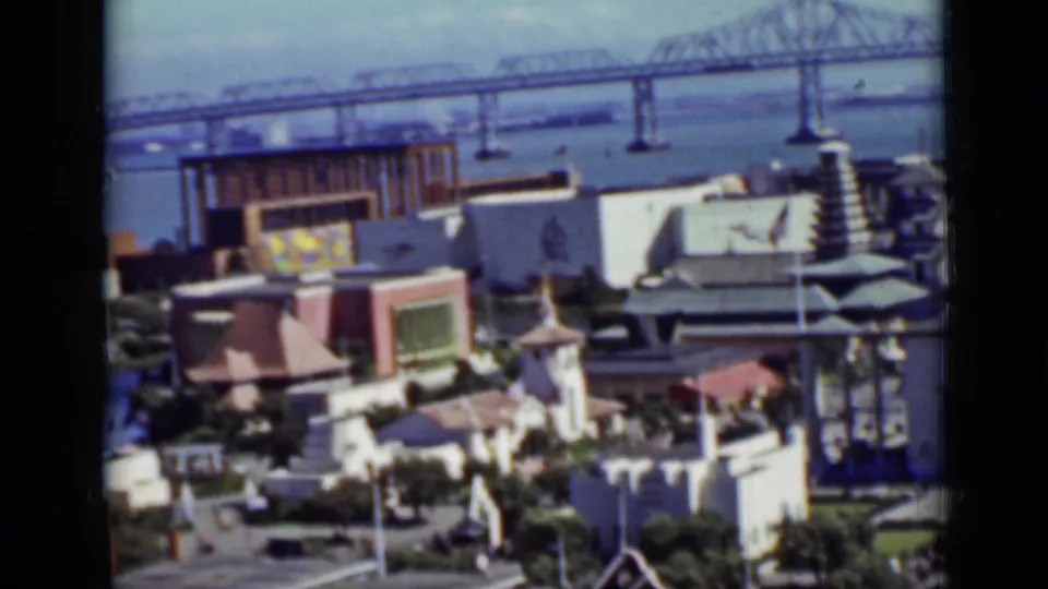 1937 San Francisco home-movie frame with a bay bridge behind colorful low buildings and landscaped paths.