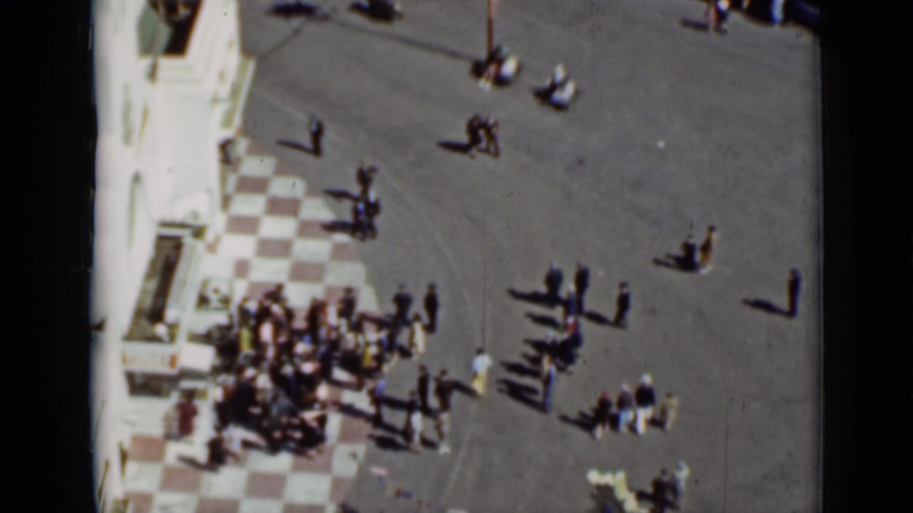 1937 San Francisco home-movie frame with groups of pedestrians crossing a broad paved plaza near a building facade.