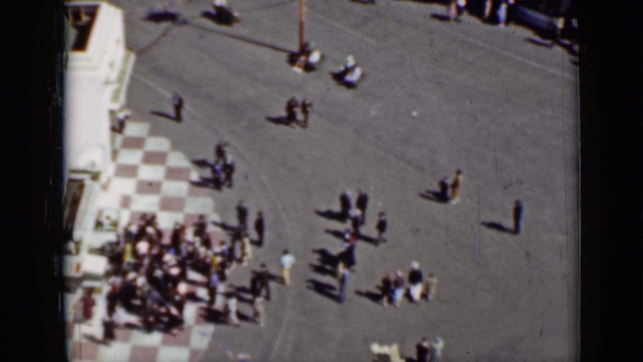 High-angle view of pedestrians crossing a paved city square near a checkered tile corner.
