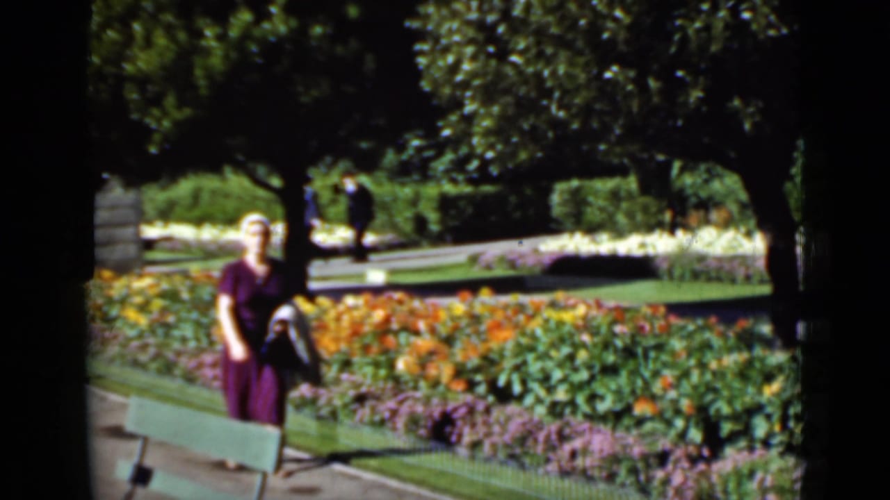 Woman in a purple dress walks past flower beds and a green bench.