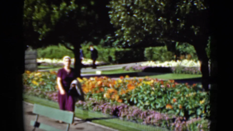 In 1937 San Francisco home-movie footage, a woman in a purple dress and white hat walks along a garden path beside bright flower beds.