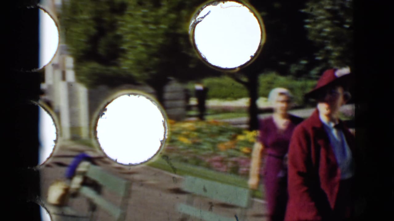 Two adult women walking on a park path beside flower beds, with bright circular film perforations at the left edge.
