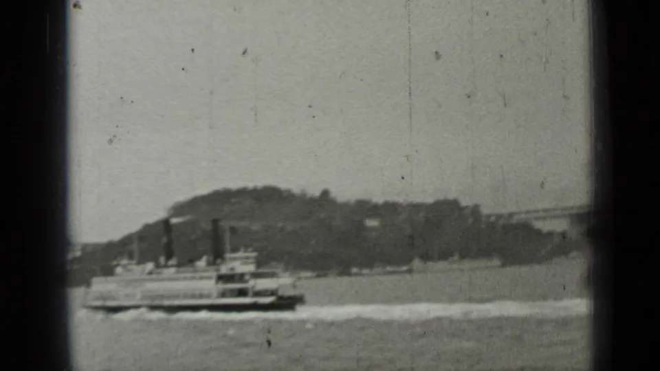 Black-and-white 1937 San Francisco waterfront frame showing a ferry crossing the bay in front of a distant hillside shoreline.