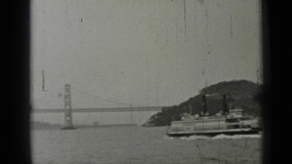 1937 San Francisco bay frame with a ferry, distant bridge tower and span, and hillside shoreline in black-and-white home-movie footage.