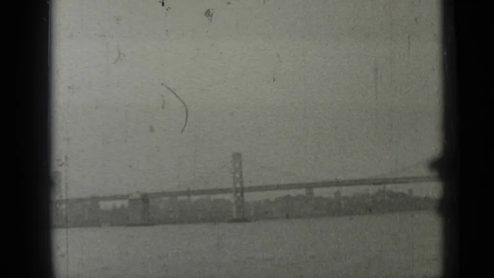 Grainy 8mm frame of the Bay Bridge stretching across San Francisco Bay, viewed from behind boat framing.