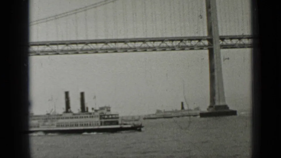 1937 San Francisco ferry passing beneath the Bay Bridge, with tower, cables, and hazy water visible in black-and-white 8mm footage.