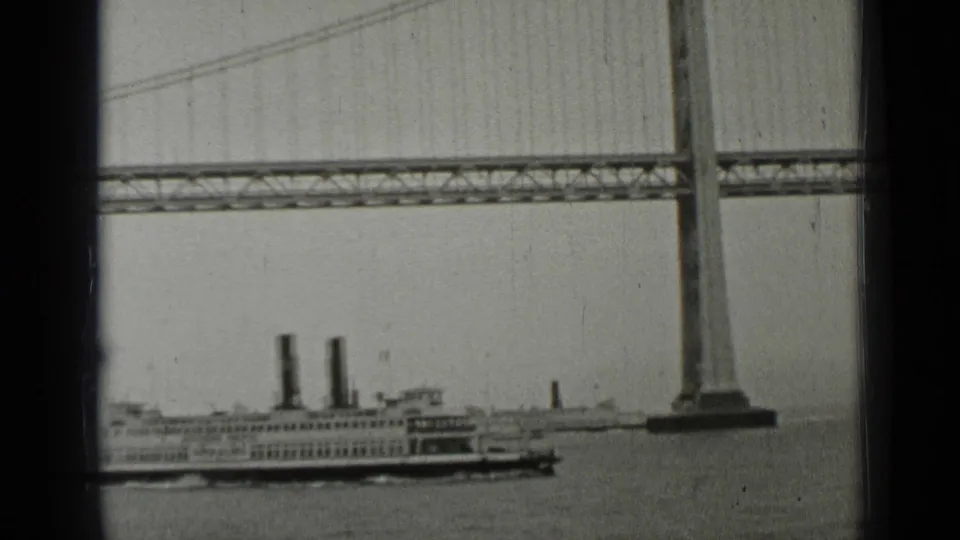 The San Francisco ferry travels under the Bay Bridge span, framed by a bridge tower and suspension cables in grainy 1937 8mm film.