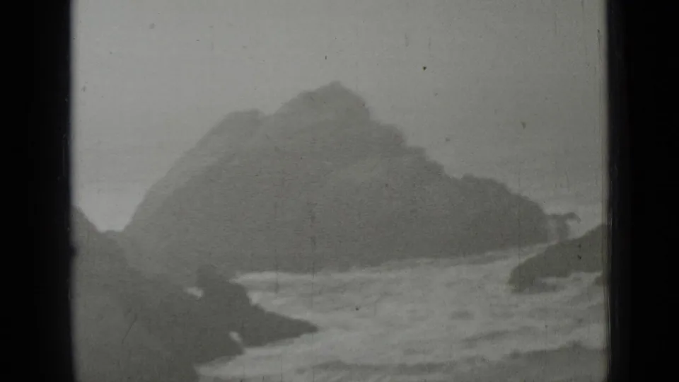 1937 home-movie frame of rugged California coast, showing surf and steep dark rocks in haze.