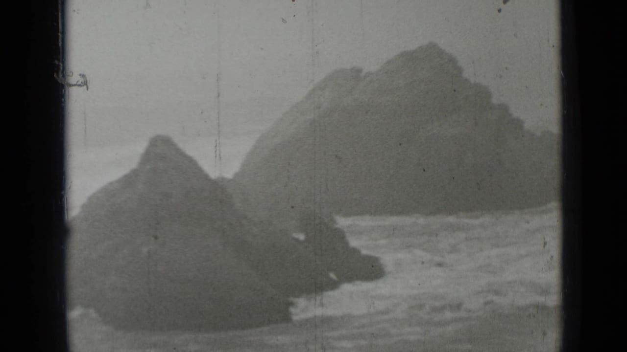 Rocky sea stacks with waves breaking around them in grainy black-and-white footage.