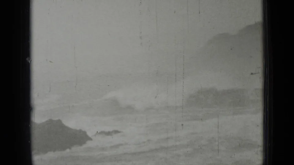 1937 8mm coastal frame with waves spreading over low rocks, a distant dark shoreline form, and heavy film wear.