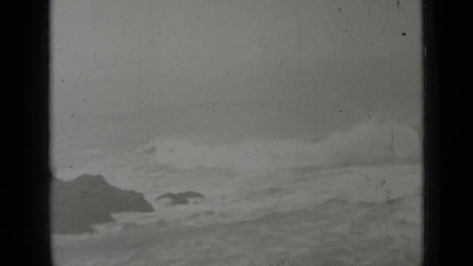 Grainy 1937 San Francisco-area coastline frame showing churning surf, low rocks, haze, and a dark rocky background.