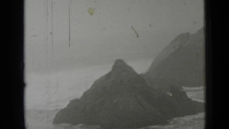 1937 coastal frame with a large dark rock in the foreground and surf breaking behind it.