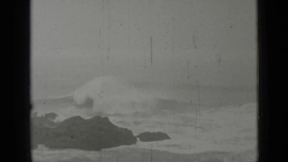 Black-and-white home-movie frame showing low coastal rocks and foamy surf in the San Francisco area.