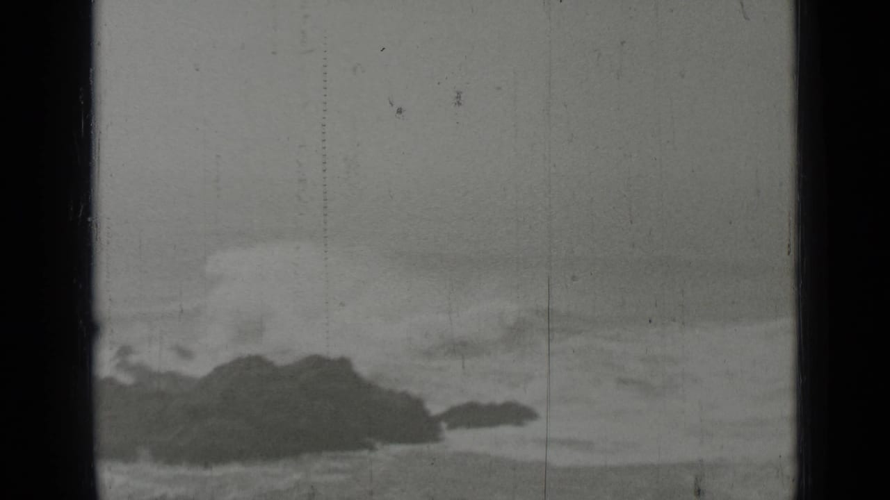 Dark rocks offshore with waves breaking under a gray sky.