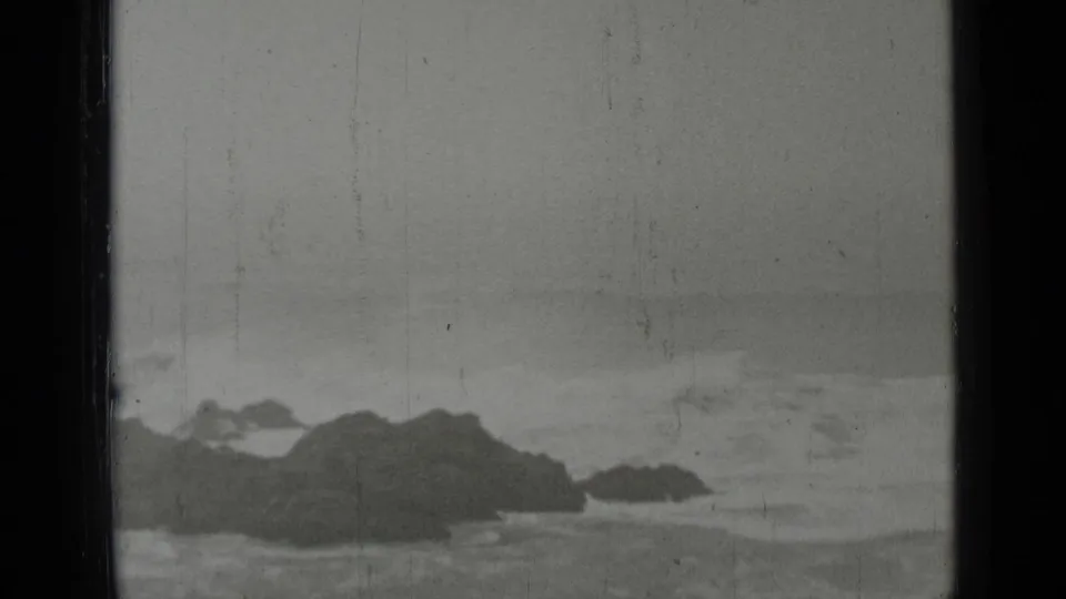 Black-and-white home-movie frame of waves striking a dark rocky outcrop along the San Francisco coast, with visible film wear.