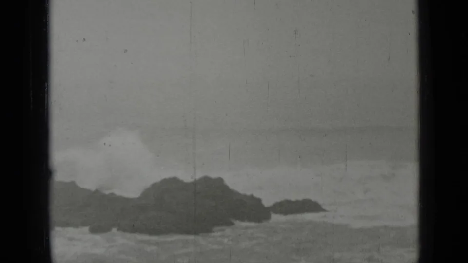 Vintage 8mm coastal frame showing foamy waves rolling toward low dark rocks, with soft focus and visible scratches.