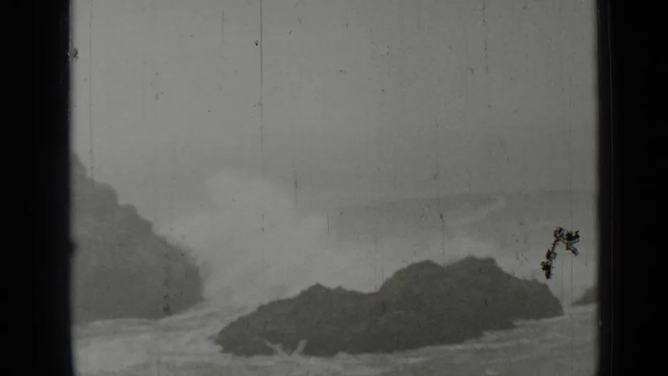 Black-and-white 8mm frame of a dark coastal rock formation bordered by pale surf near San Francisco, with dust and scratches visible.