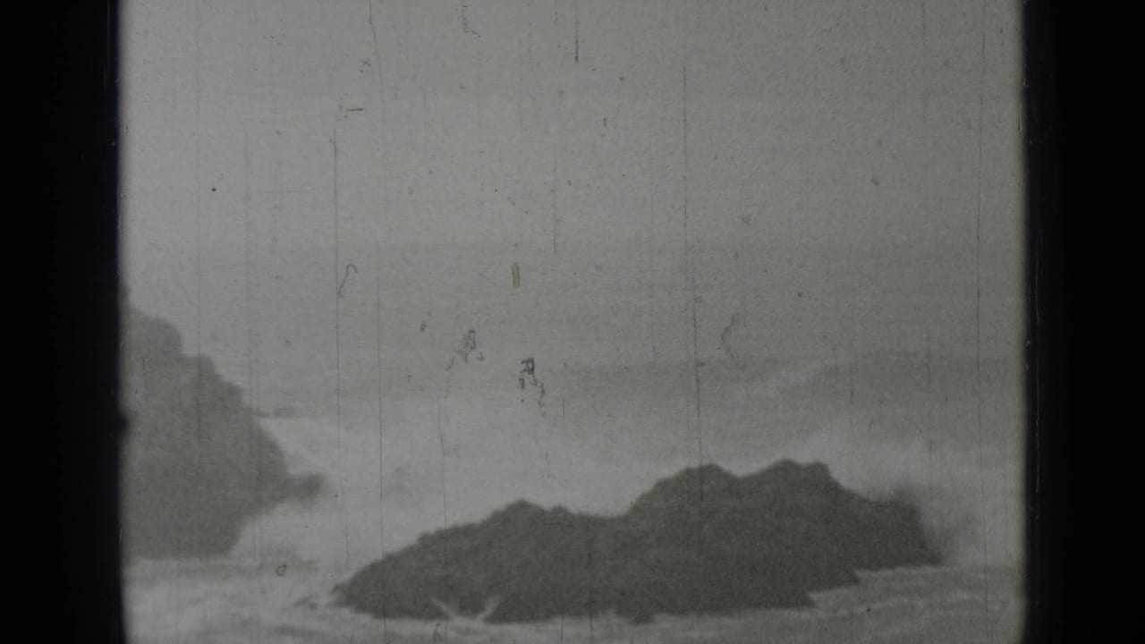 Dark rocks sit offshore as waves break under a hazy sky.