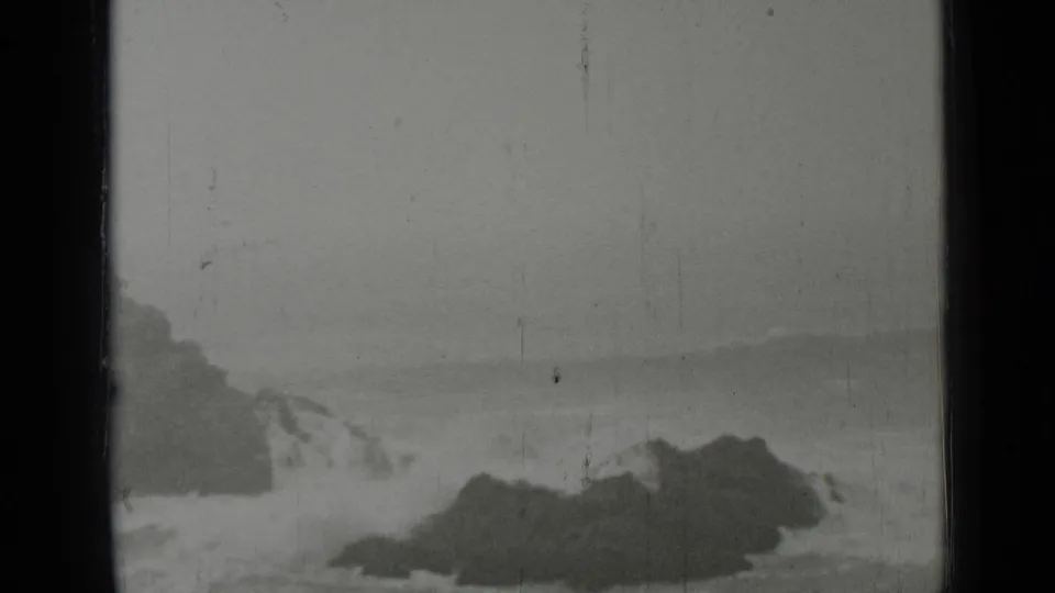 Black-and-white 8mm view of San Francisco-area rocks with turbulent surf and a soft, misty horizon line.