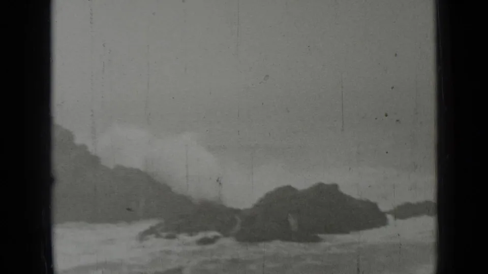 1937 coastal frame showing rough water moving around dark rocks, with visible 8mm grain and wear marks.