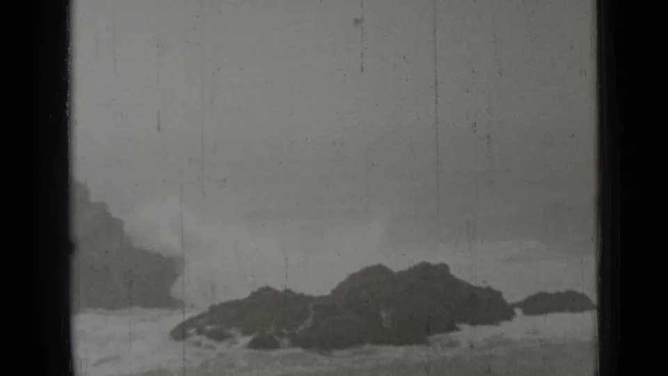 San Francisco coastal home-movie frame from 1937 with low dark rocks, pale surf, foggy background, and black-and-white film texture.
