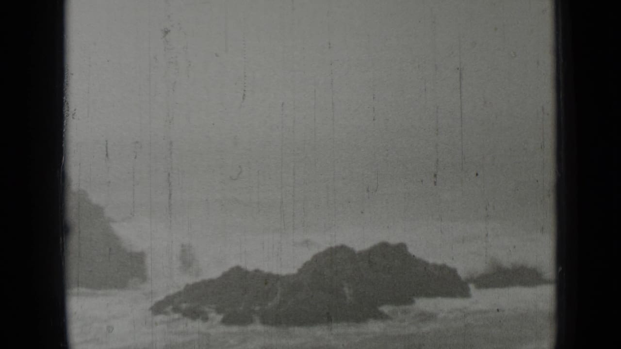White surf breaks around dark rocks under a gray sky, framed by black 8mm film edges.