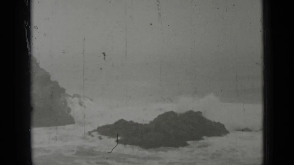 1937 San Francisco 8mm frame showing a low dark rock formation with white surf rolling around it.