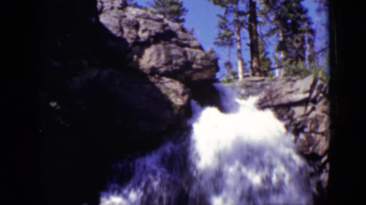 White water pours from a dark rock opening beneath evergreen trees.
