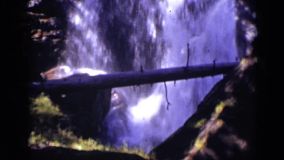 Colorado home-movie frame from 1938 showing a bright cascade, foreground log rail, and shadowed rock walls.