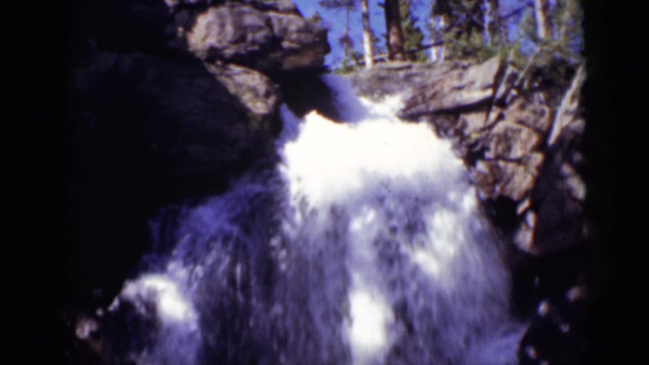 Rocky waterfall spilling over dark stone with blue sky and trees above.