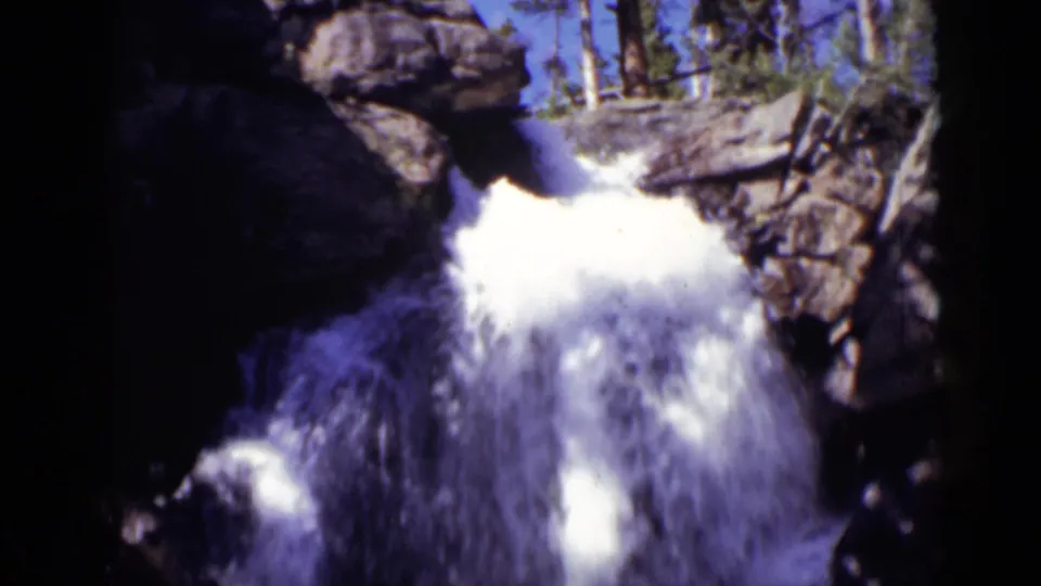 1938 Colorado 8mm frame of bright white waterfall spray dropping through dark rocks beneath trees and blue sky.
