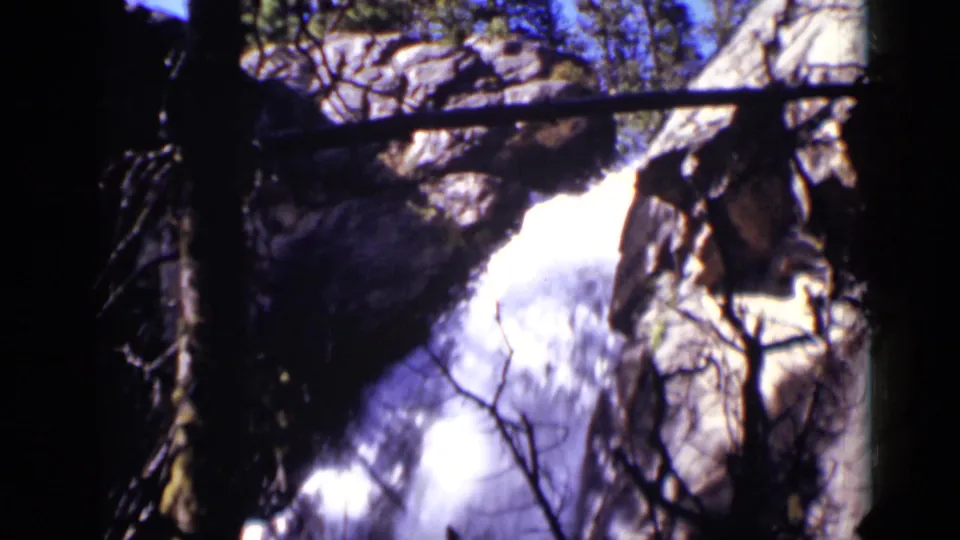 Colorado 1938 home-movie frame of a waterfall partly obscured by branches, rocks, and a horizontal fallen tree.