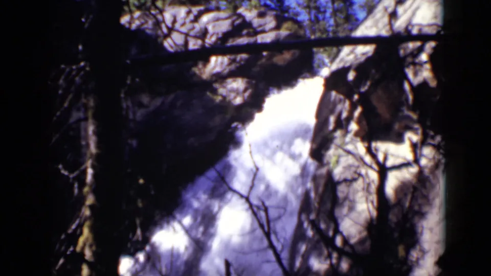 Colorado forest view with a white waterfall dropping beside rock faces, framed by dark branches in 8mm home-movie color.