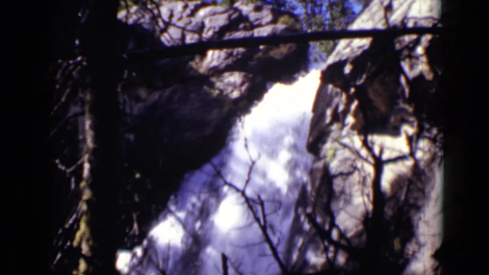 8mm frame of a Colorado waterfall descending through rocks, with foreground branches and strong contrast from the original film exposure.