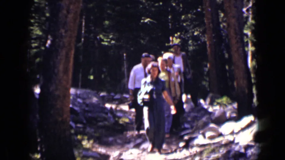 1938 Colorado home-movie frame showing a small group walking along a sunlit forest trail between tall trees and scattered rocks.