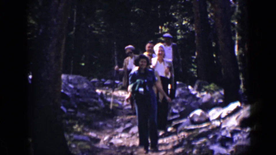 1938 Colorado home-movie frame of adults hiking through forest, including a front hiker with a camera at her chest.