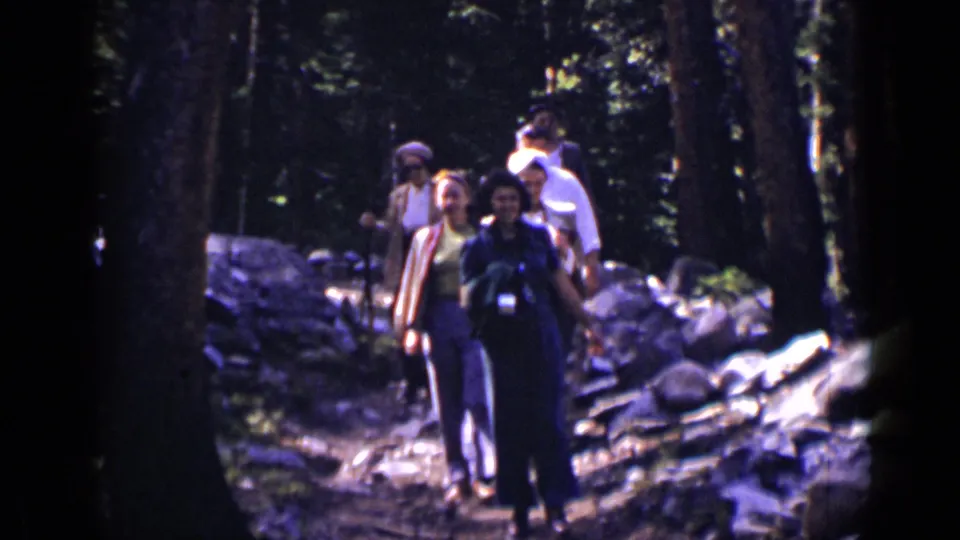 Adult hikers in 1938 Colorado forest footage pass near the camera, with rocks, tall trees, and walking sticks visible.