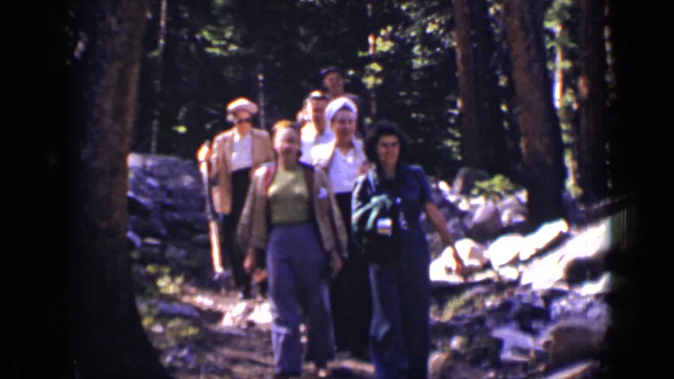 Several hikers in 1930s attire walk toward the lens on a Colorado forest trail with rocks and bright sun patches.
