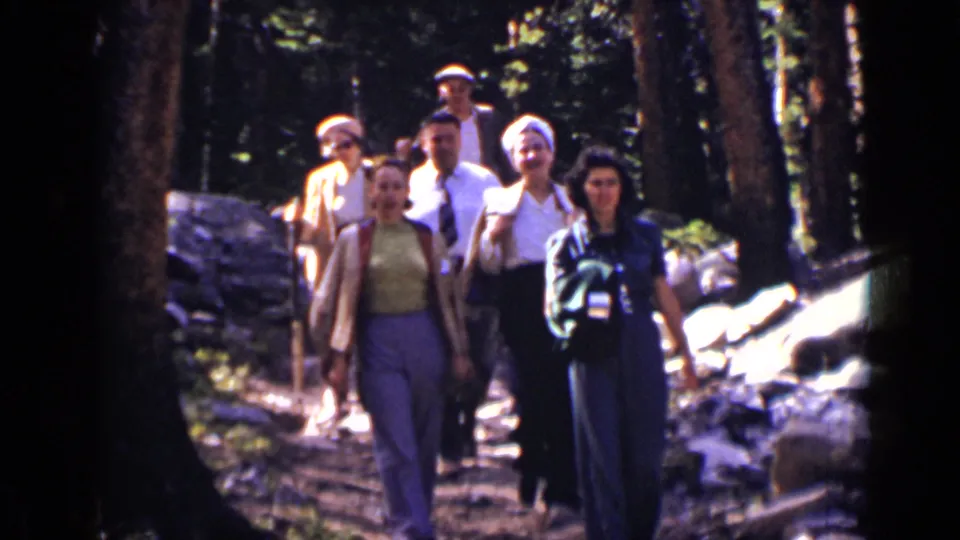 The hiking group appears closer to camera on the wooded Colorado trail, with faces, hats, jackets, and blouses visible.