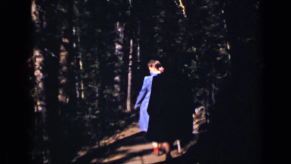 In 1938 Colorado home-movie footage, a woman in a blue dress and red shoes walks away along a sunlit wooded path.