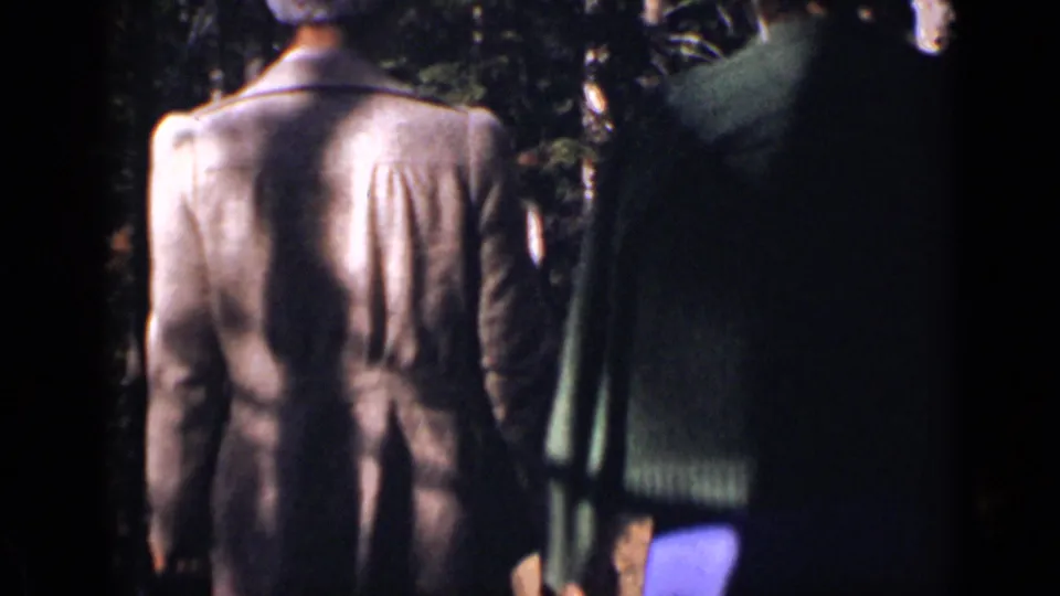 Two adults in a light jacket and green sweater walk away along a shadowed Colorado wooded path in 1938 8mm footage.