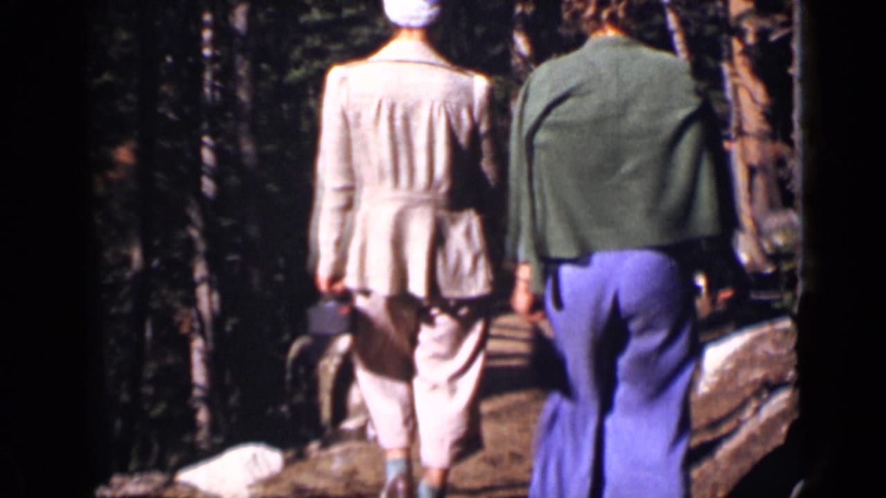 Two people continue along a dirt trail among trees and rocks.
