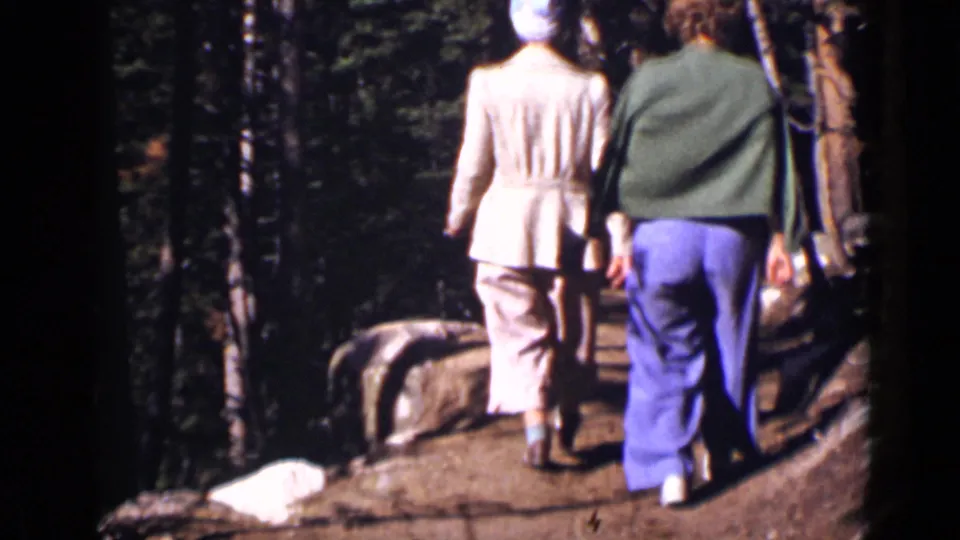 Two adults, one in a light outfit and one in blue trousers, walk deeper into a Colorado wooded trail scene.