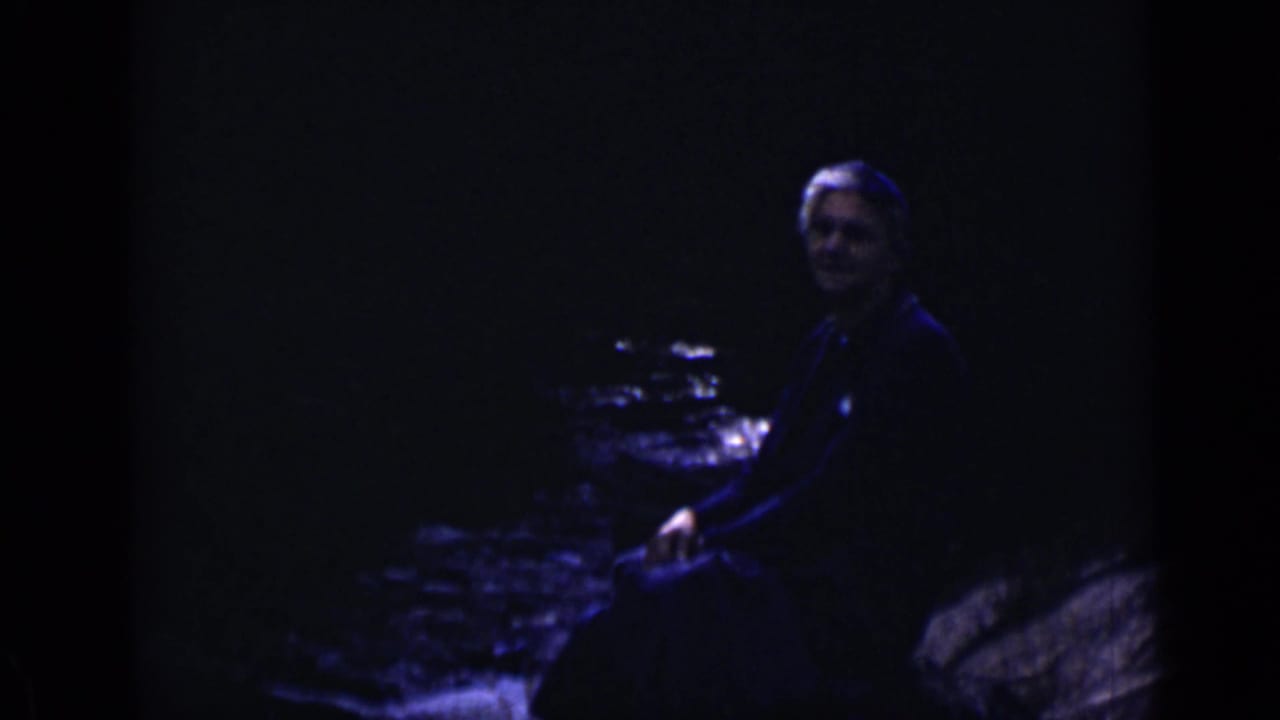 Adult seated on rocks beside dark flowing water.
