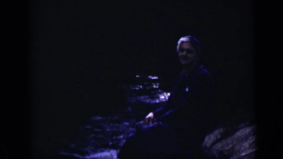 1938 Colorado 8mm frame showing an adult in dark clothing seated on a slanted rock beside a shaded river.