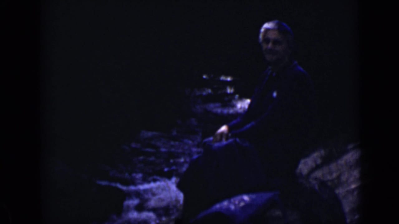 1938 Colorado home-movie frame of an adult looking toward the camera while seated beside a rocky riverbank.