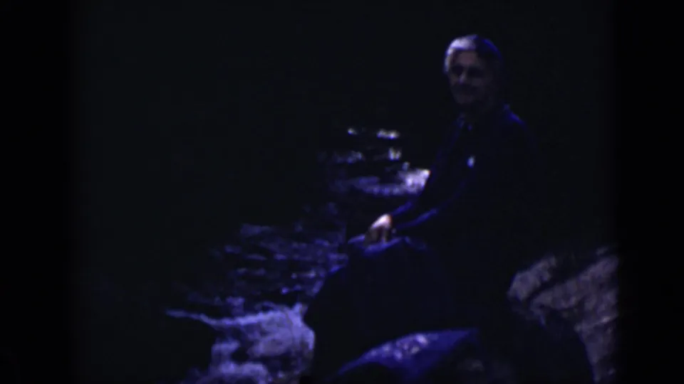 1938 Colorado home-movie frame of an adult looking toward the camera while seated beside a rocky riverbank.